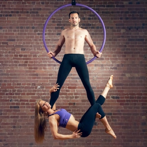 Cerceau aérien violet X-Pole devant un mur de briques avec un modèle masculin dessus, auquel une modèle féminine est accrochée par les jambes